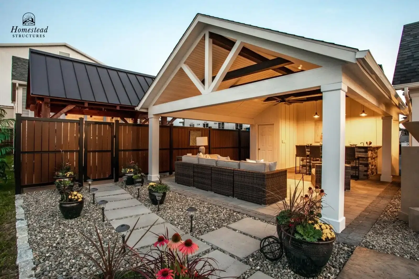 A backyard patio with a covered outdoor living space featuring a wicker sectional sofa, a bar with high chairs, and decorative potted plants, surrounded by a gravel yard with stepping stones and a wooden privacy fence.