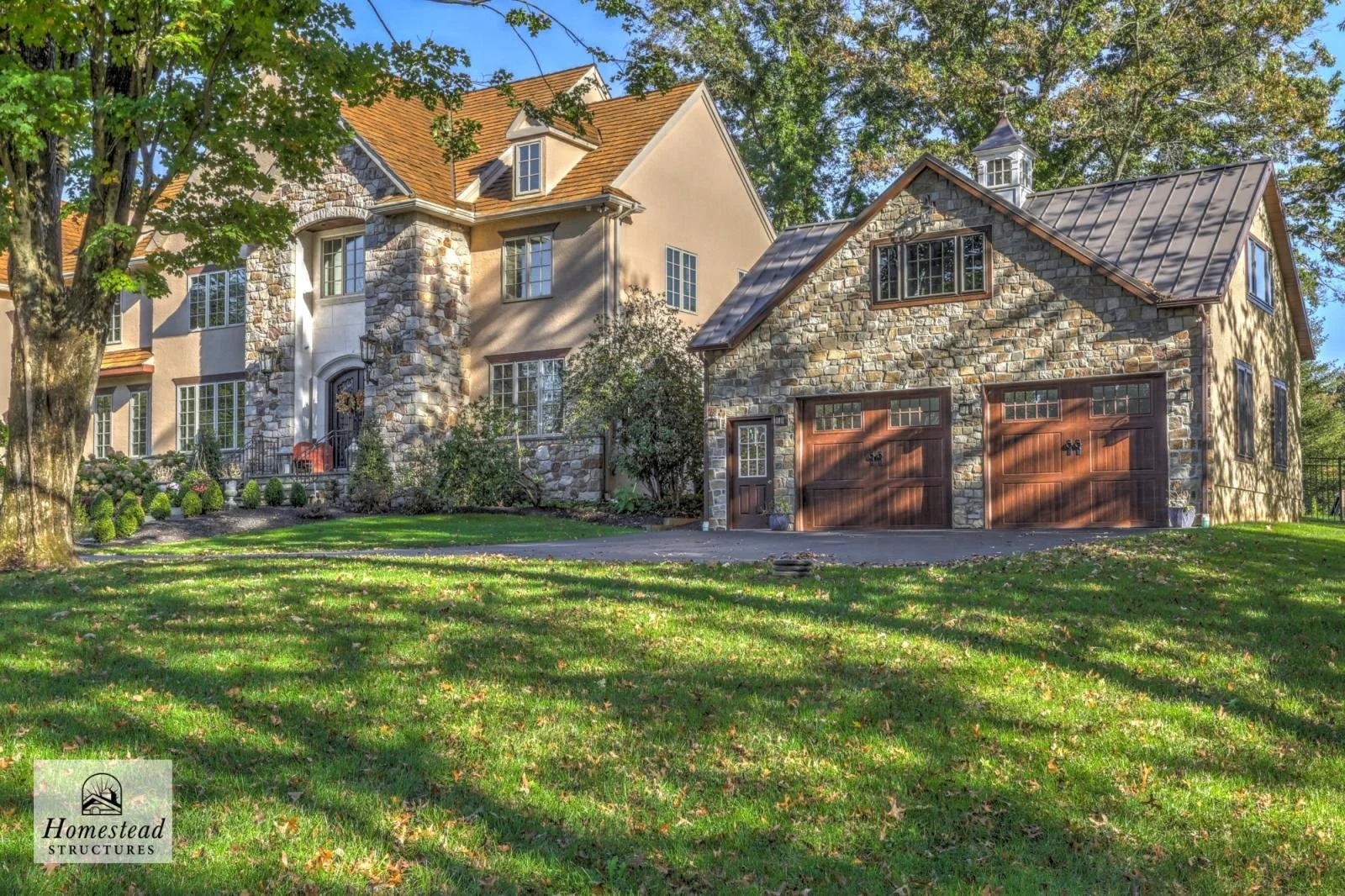 A large house with a stucco and stone exterior, and a two-car garage with wooden doors, surrounded by a lush green lawn and trees.