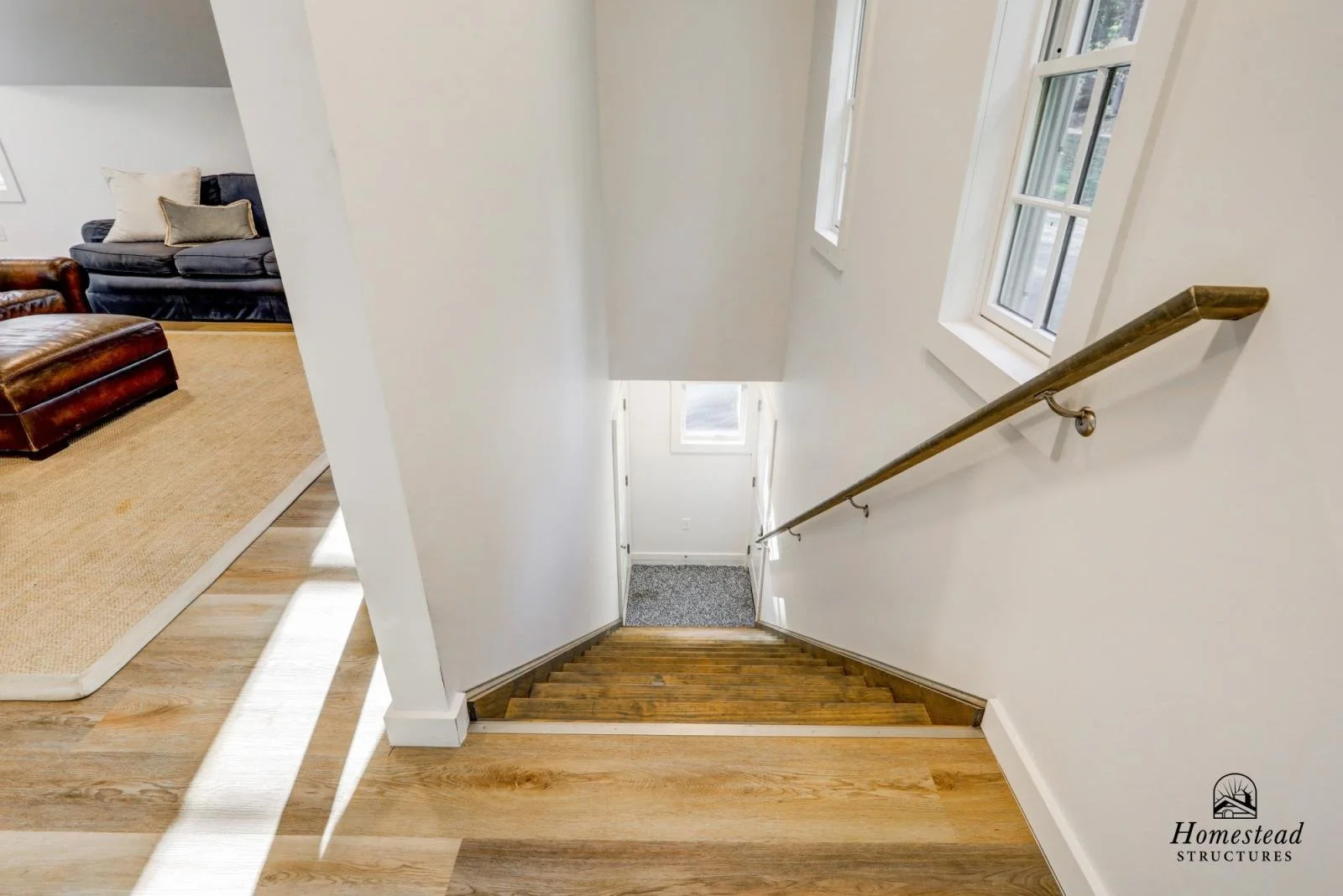View of a staircase with wooden steps and a handrail, leading down to a lower level with carpet, in a bright home interior. Part of a living room with a leather and fabric sofa, a tan rug, and hardwood flooring is visible to the left.