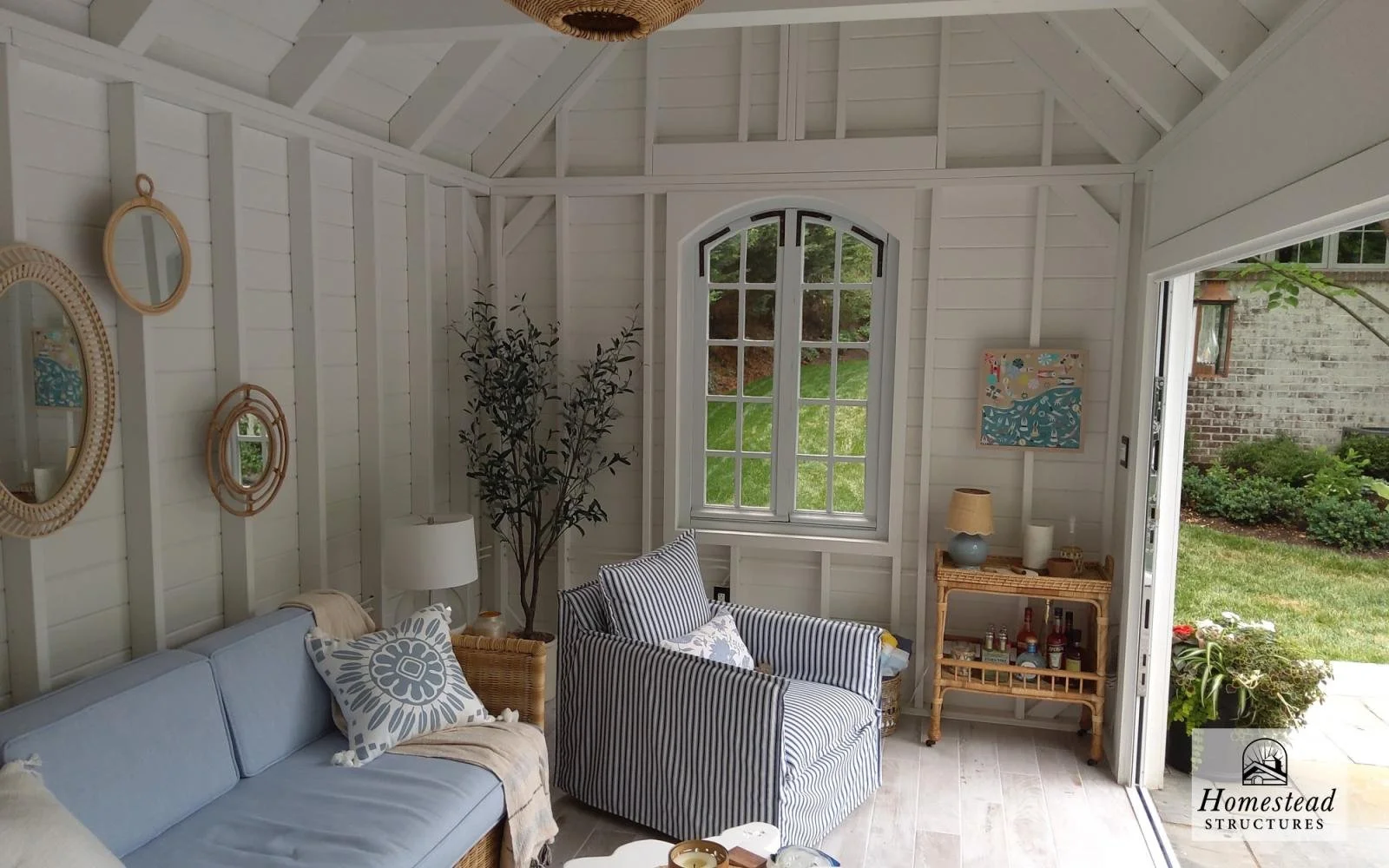 Interior of a sunroom with white wooden walls and ceiling, featuring a light blue loveseat with patterned pillows, a striped armchair, a wicker side table, a lamp, and barware, with a large window and an open door showing a grassy garden outside.