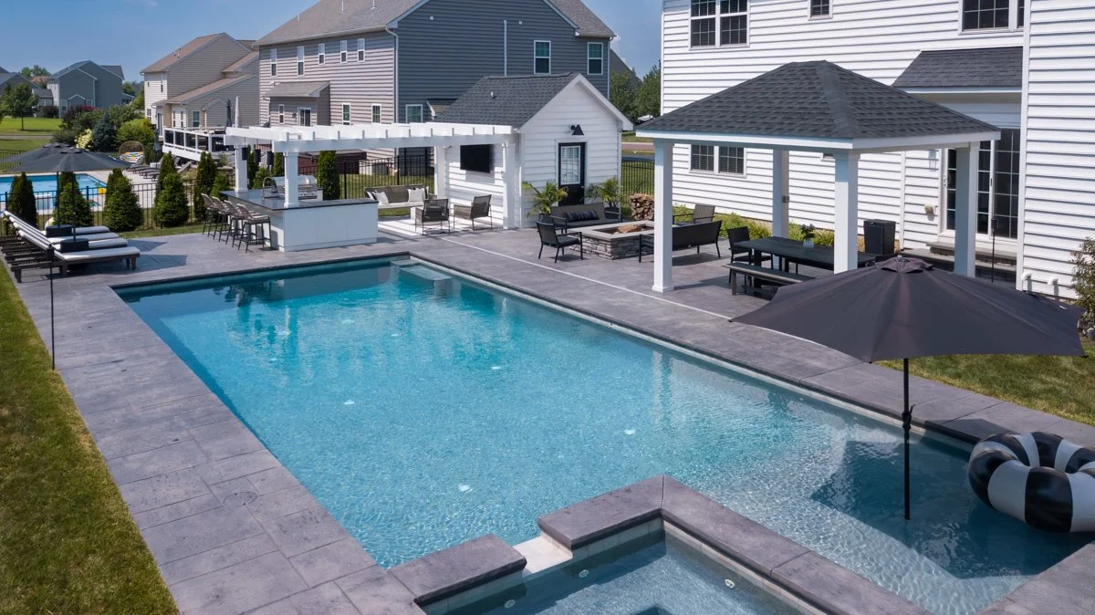 Backyard view with a rectangular swimming pool, poolside lounge chairs, a shaded sitting area with outdoor furniture, a bar area with barstools, a pool float, and a black umbrella, surrounded by houses and a well-maintained lawn.