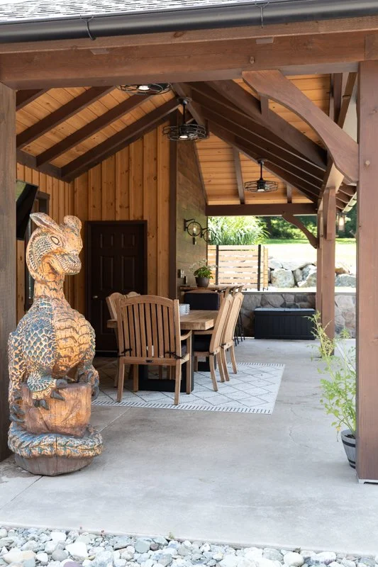 Covered patio with wooden beams and ceiling, outdoor dining table with six chairs, decorative lizard sculpture, potted plant, bench, and view of lush green landscape in the background.