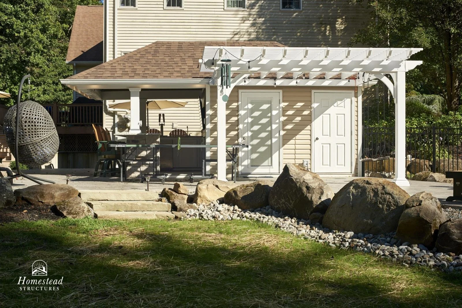 Backyard patio with a white pergola, outdoor furniture, a hanging egg chair, a grill, and large rocks, with a house and trees in the background.