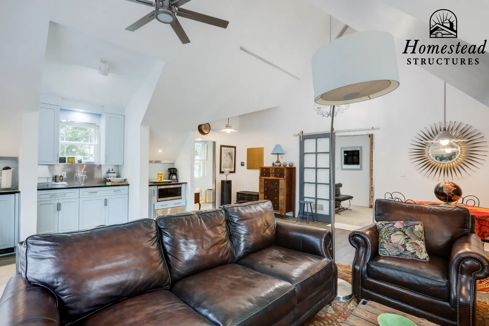 Living room with dark brown leather sofas, a standing lamp, a decorative mirror, and a ceiling fan. The space includes a kitchen area with white cabinets and a window, with various furniture and decor items visible.
