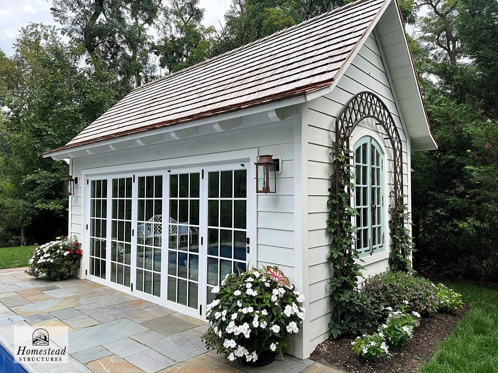 Small white garden shed with front sliding glass doors, a gable roof with brown shingles, and decorative black metal arch over a window on the side, surrounded by plants and flowers, with a stone patio in front.