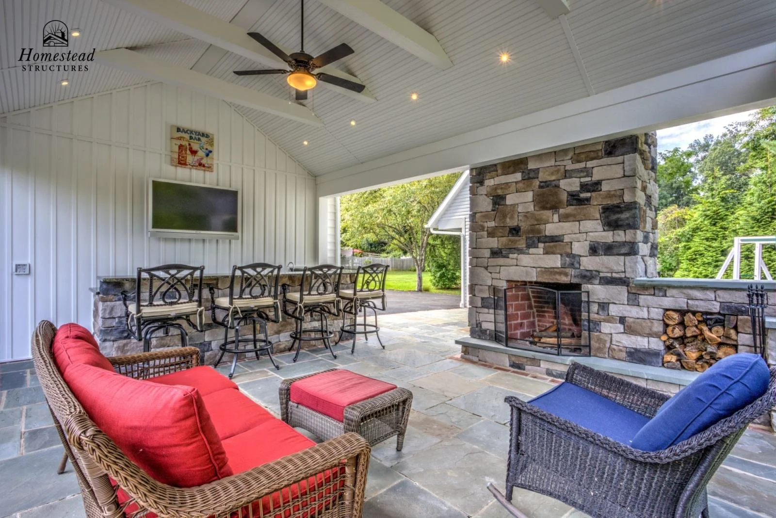 Covered outdoor patio with a stone and brick fireplace, wicker and metal seating, a ceiling fan, and a view of greenery outside.
