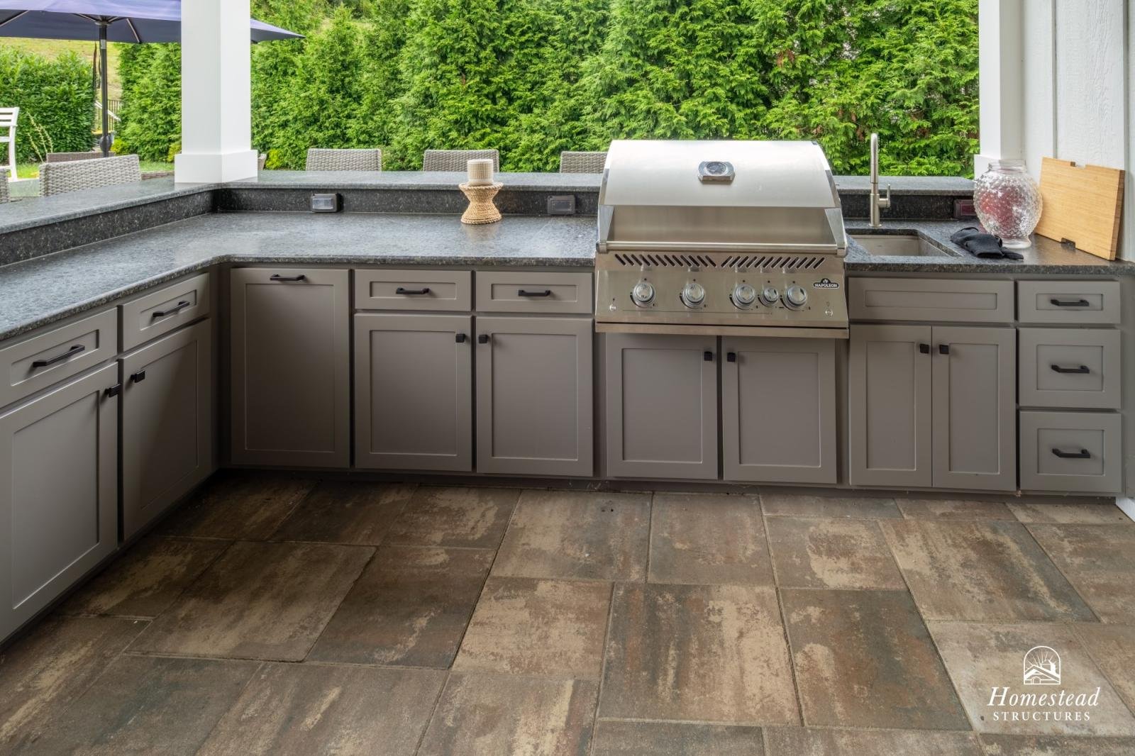 Outdoor kitchen with gray cabinets, a metal grill, and a granite countertop, overlooking a green leafy background.