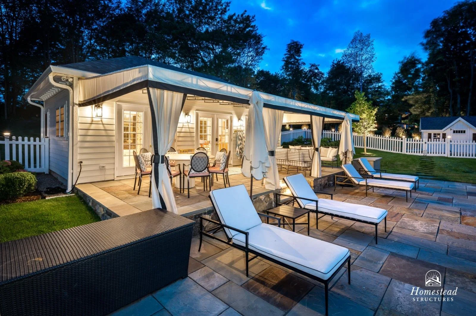 A backyard patio at dusk with lounge chairs, a dining table with chairs under a covered area with curtains, and a white fence surrounding the yard, decorated with outdoor furniture and illuminated by lights.