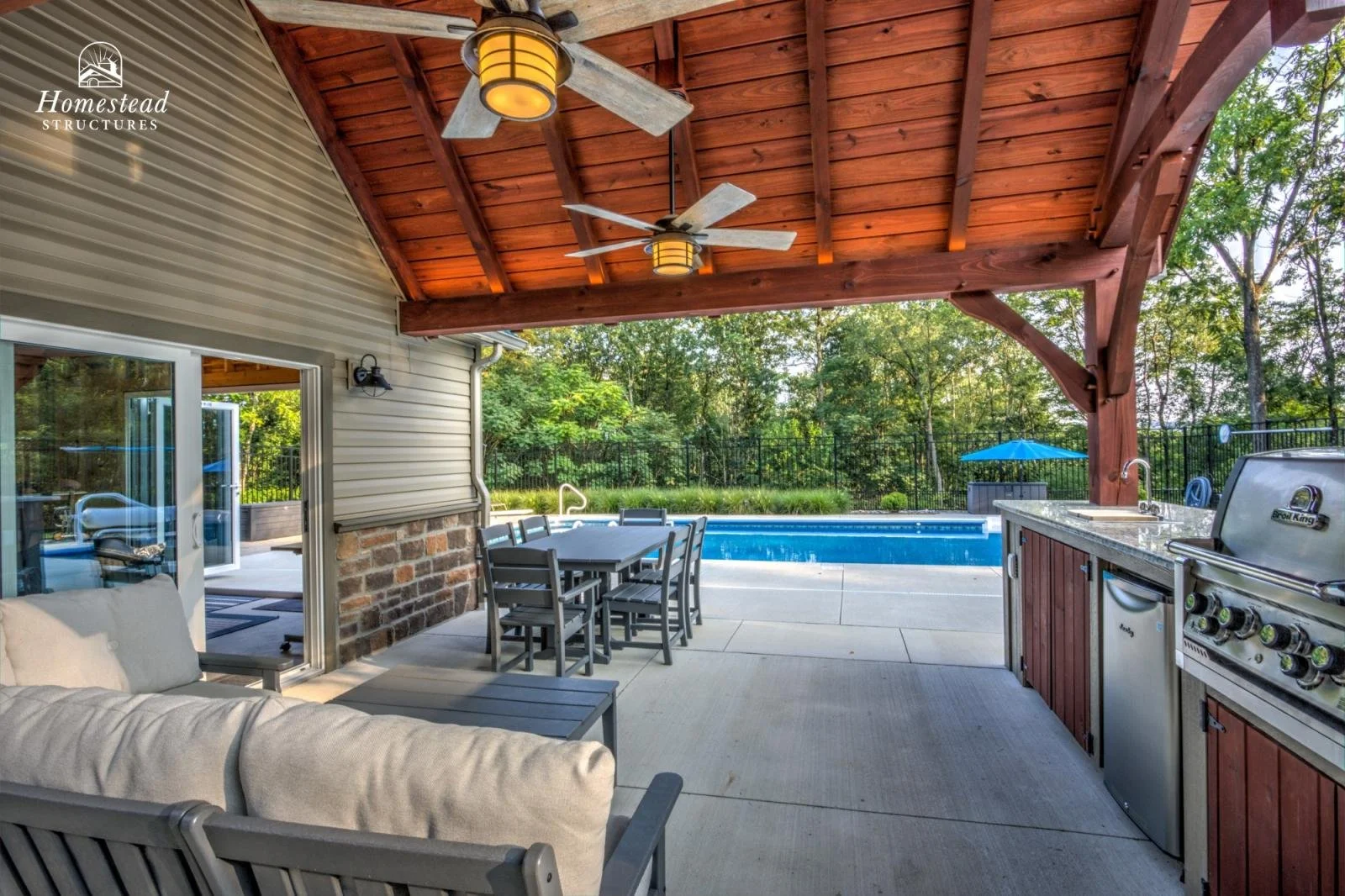 Covered outdoor patio with ceiling fans, a sofa, dining table with chairs, a built-in grill, a small refrigerator, a sink, and a swimming pool in the background surrounded by trees.