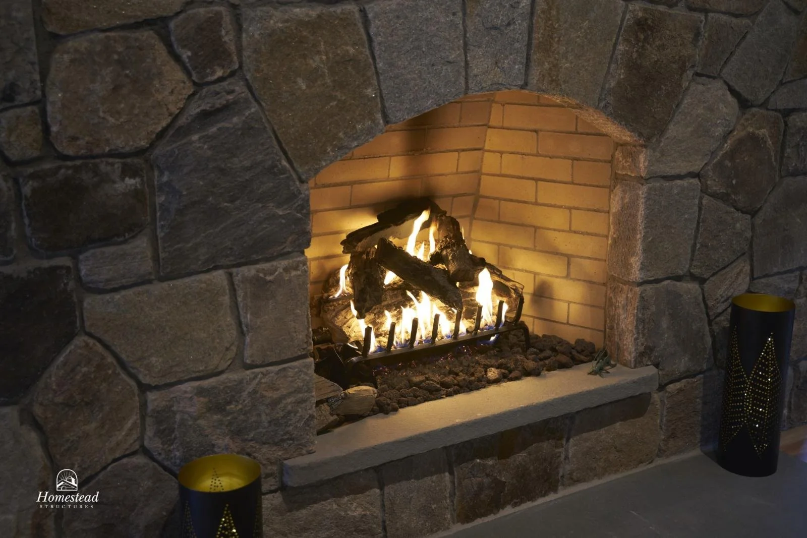 A stone fireplace with a roaring fire inside, surrounded by a brick interior. Two black candles with gold designs are placed on the hearth, and a small logo with the text 'Homestead Structures' is visible in the bottom left corner.