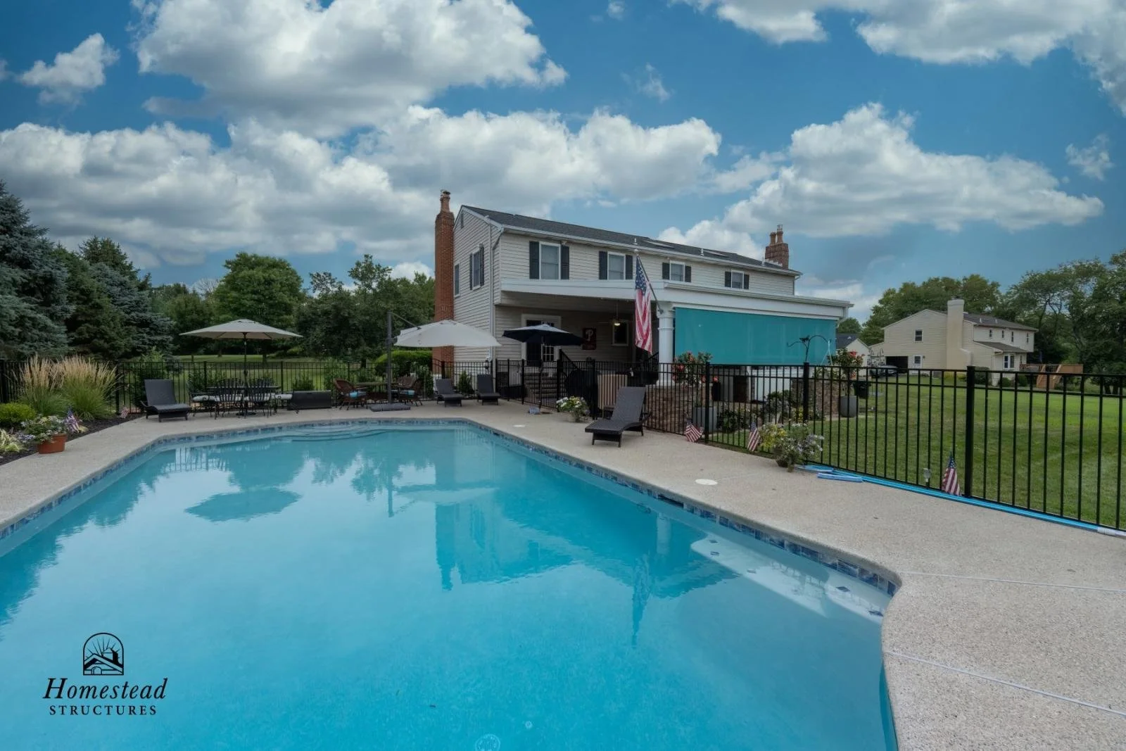 A backyard with a swimming pool, surrounded by patio furniture, umbrellas, and a black fence. In the background, a white two-story house with a chimney, American flag, and green lawn, under a partly cloudy sky.
