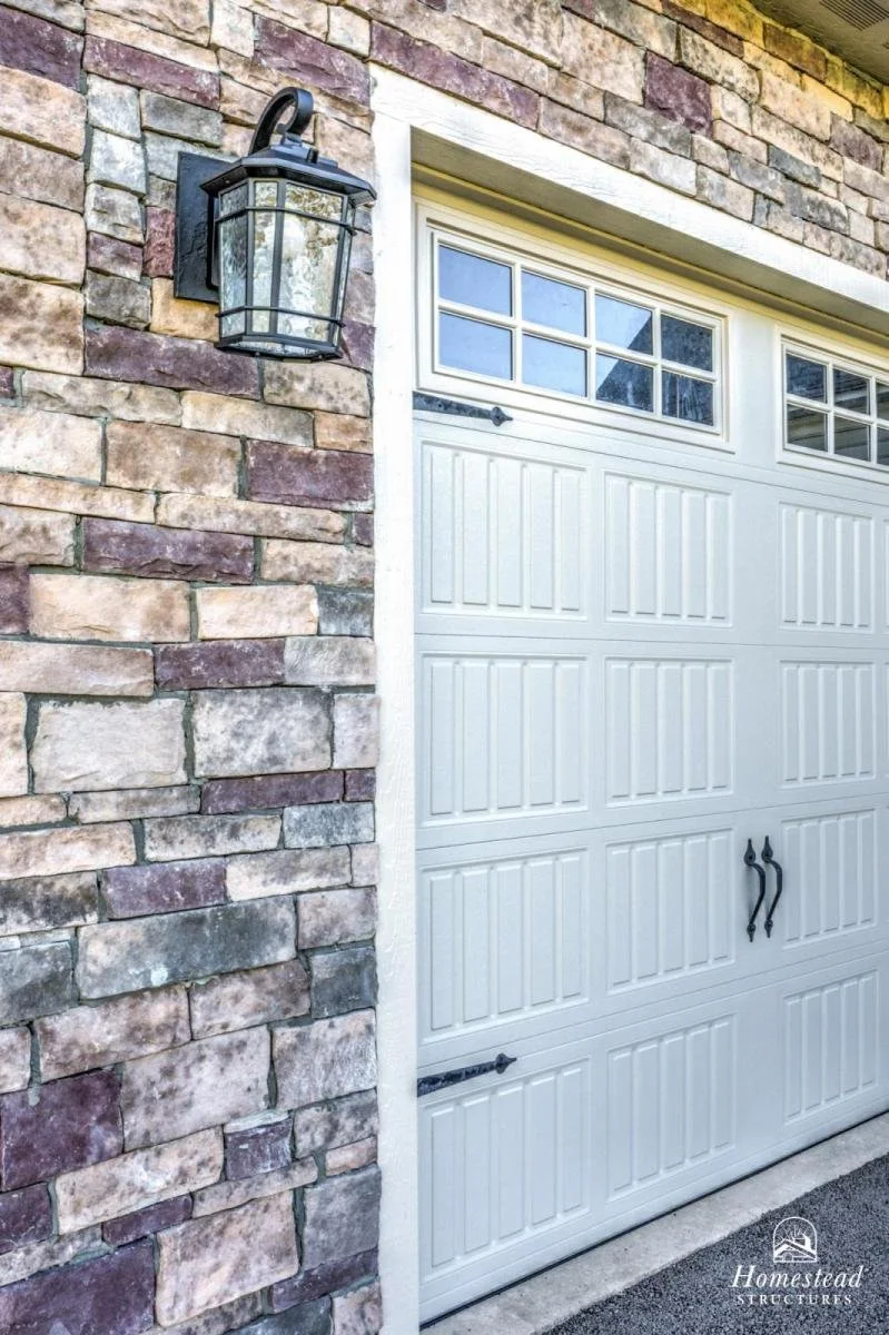 Exterior view of a white garage door with windows at the top, a brick wall to the side, and an outdoor lantern above the door.