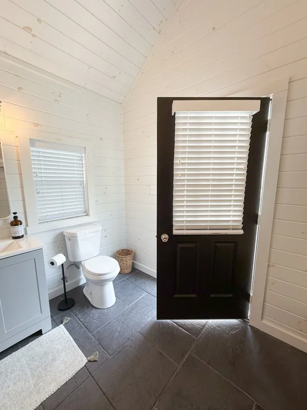 A small bathroom with white wooden walls and ceiling. There is a black door with a window covered by white blinds. Inside, a white toilet with a wicker basket beside it, a gray cabinet with a soap dispenser and a mirror above it, and a window with wh
