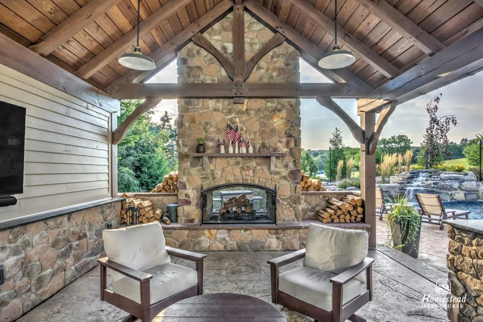 Covered outdoor patio with stone fireplace, stacked firewood, two light-colored armchairs, and a pool with lounge chairs in the background.