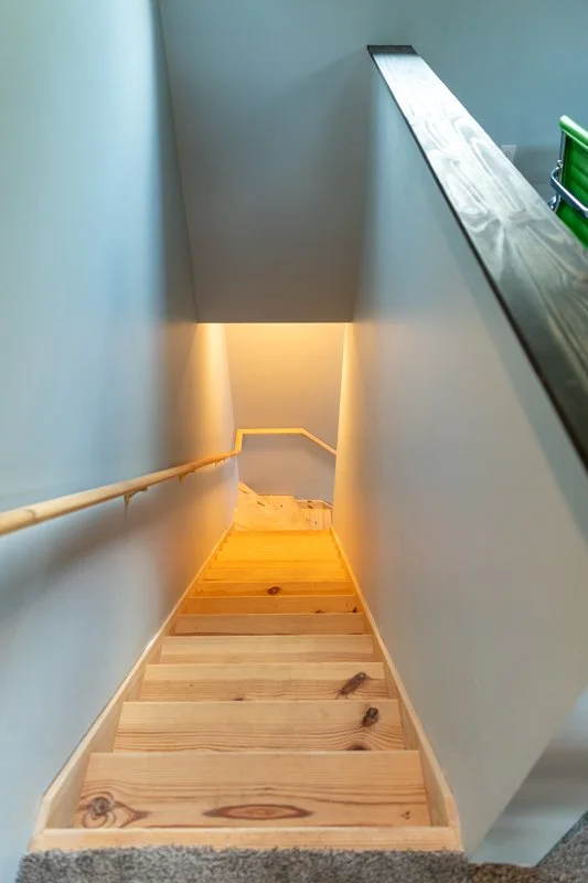 Wooden staircase leading down to a lower level, with a beige wall and a handrail on the left side and a gray wall with a black railing on the right side.