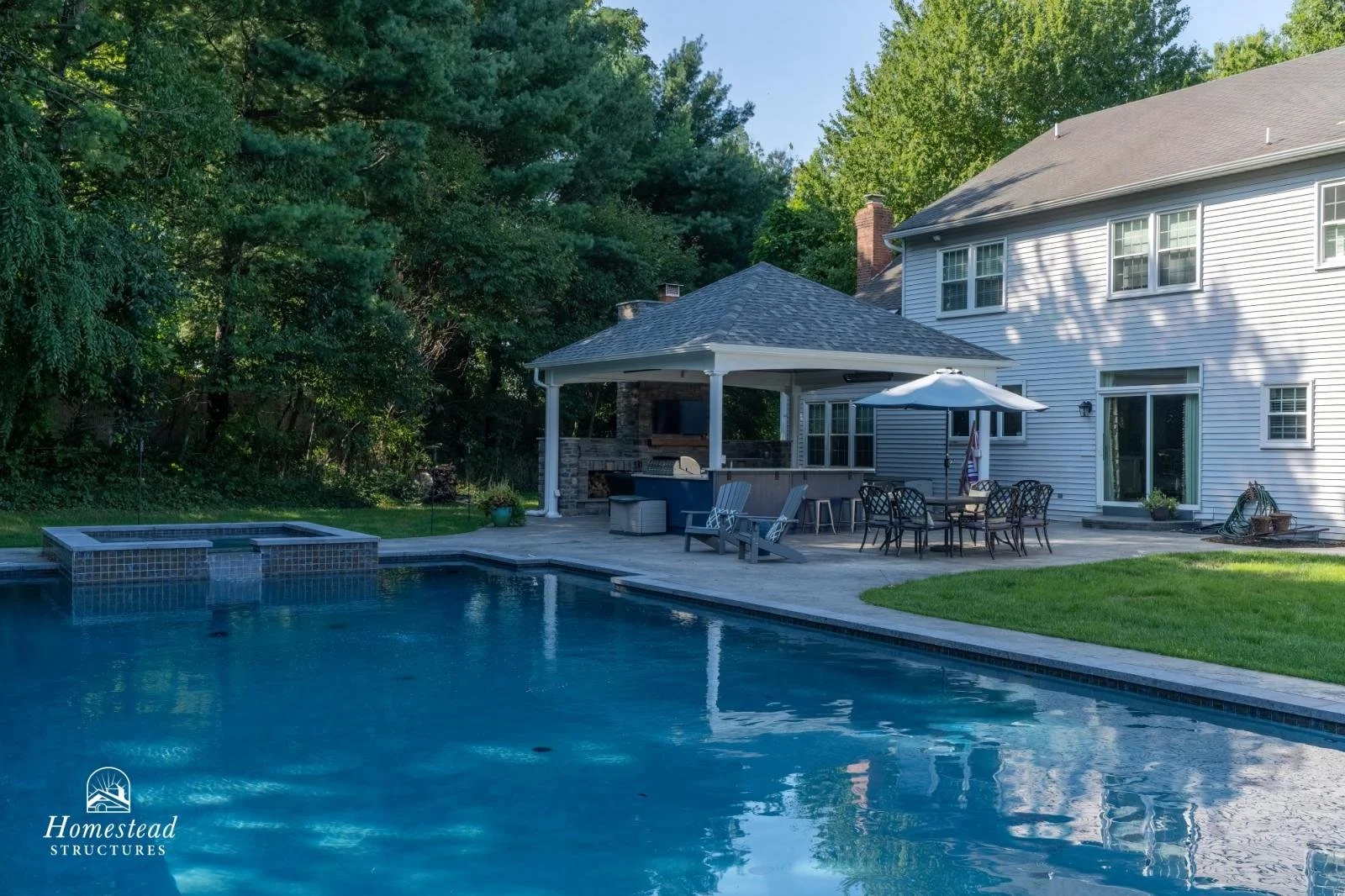Backyard with a swimming pool, patio furniture, a gazebo with a built-in fireplace, and a white two-story house with sliding glass doors and windows, surrounded by green trees.