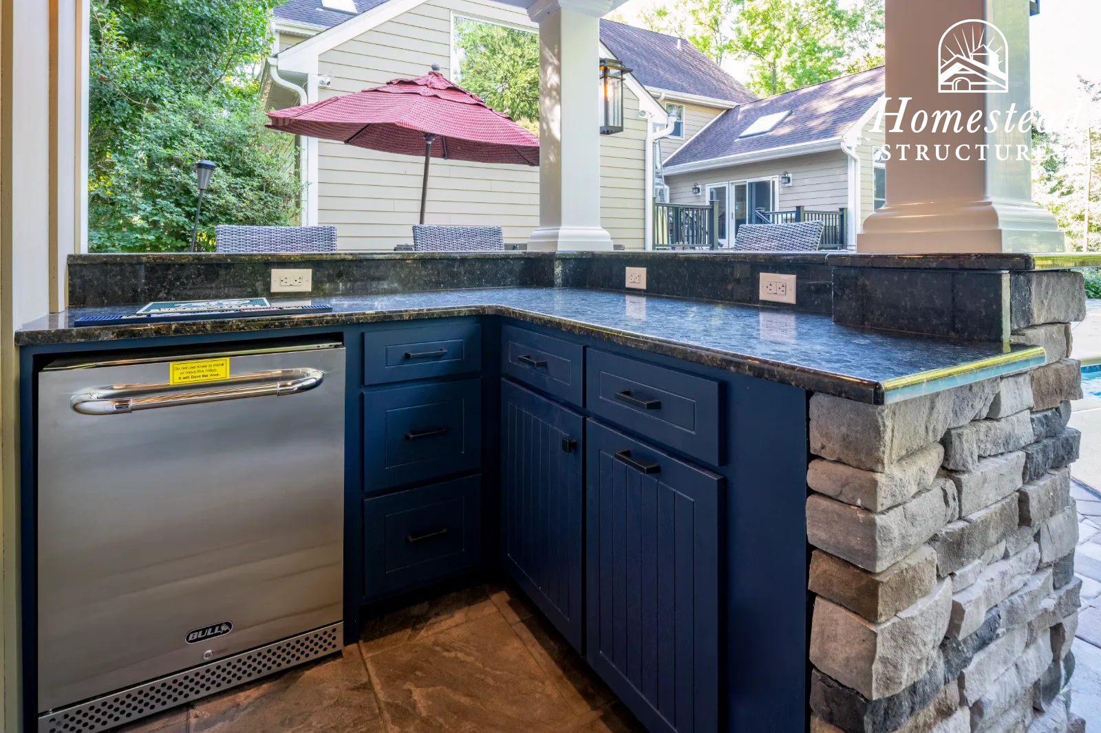 An outdoor kitchen with a stone base, dark blue cabinetry, a black granite countertop, and a built-in small stainless steel refrigerator. The kitchen is adjacent to a patio with outdoor chairs and a red umbrella, and residential houses are visible in