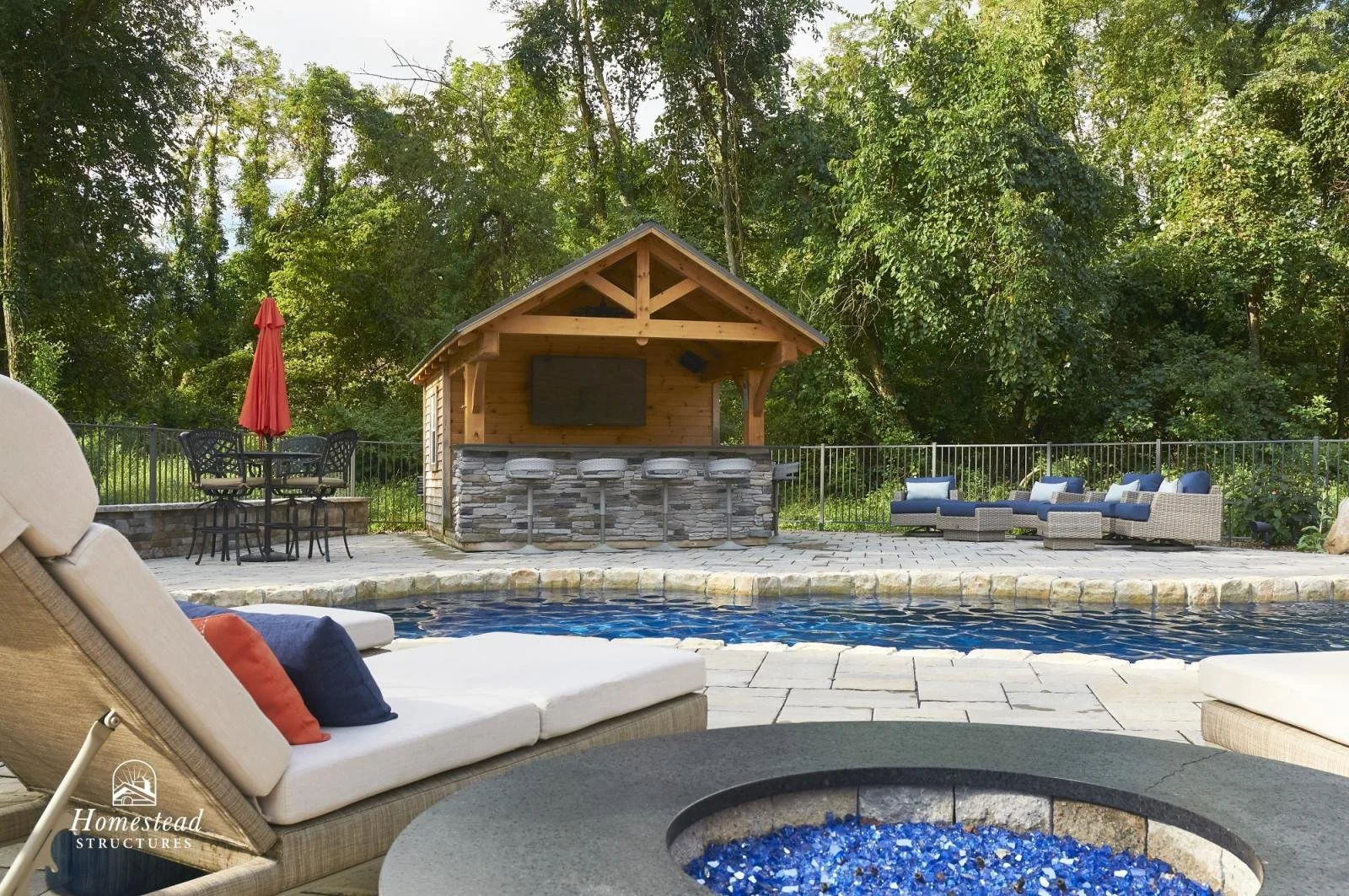 Outdoor backyard with a swimming pool, lounge chairs, a fire pit with blue glass, a bar area with barstools, and a covered outdoor kitchen with a TV, surrounded by trees.