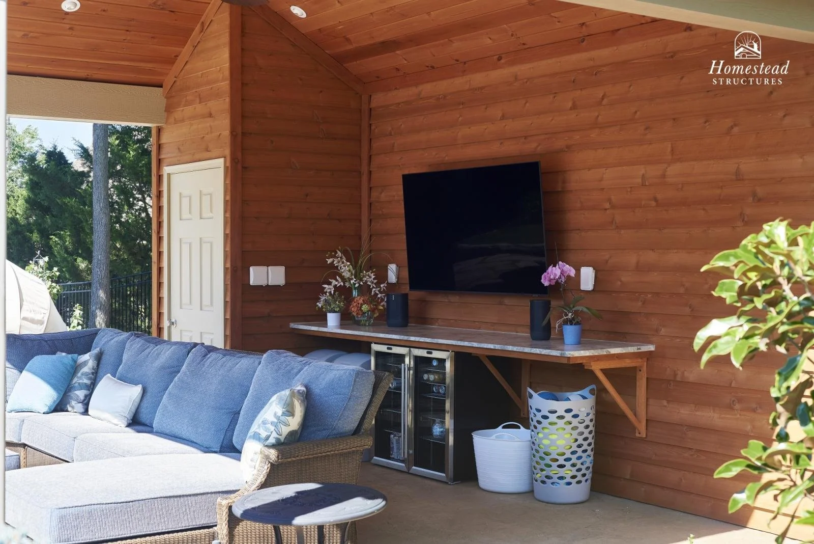 Outdoor living space with blue cushioned sectional sofa, decorative pillows, a small side table, a wall-mounted television, a marble-topped shelf with potted plants, a mini fridge, and laundry baskets against a wooden wall with a door and windows sho