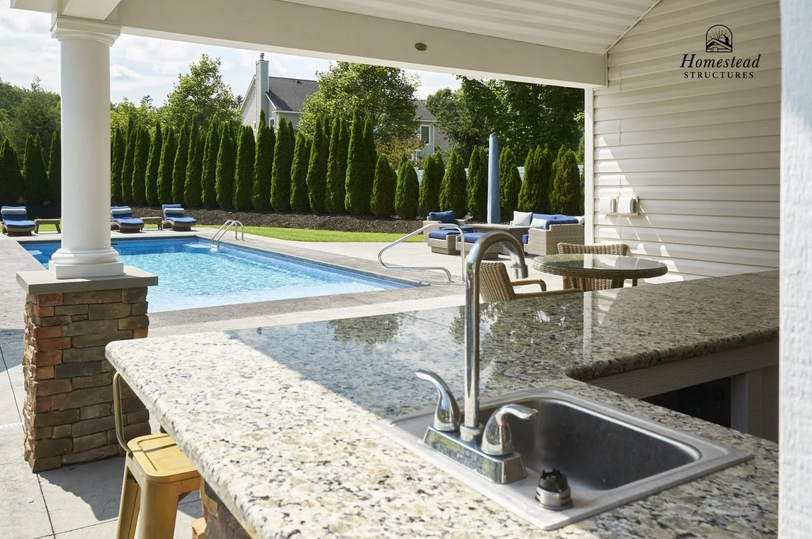 View of an outdoor swimming pool and patio area from a kitchen with granite countertops, including a sink and stool, in a backyard with tall green bushes and several lounge chairs.