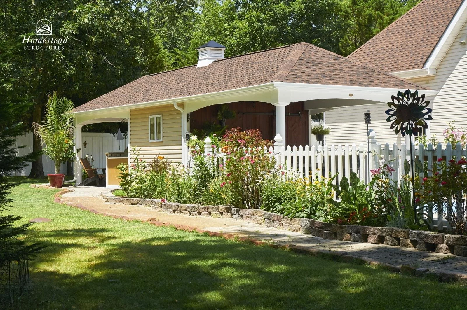 A backyard garden with a brick pathway, white picket fence, blooming flowers, and a small building with beige siding and a brown shingle roof.