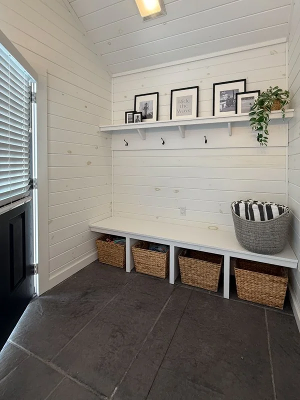 A small entryway nook with white paneled walls and ceiling, dark gray tiled floor, and a bench with four storage baskets underneath. A white shelf with framed pictures, a sign, and a plant is mounted on the wall above the bench. There are four hooks 