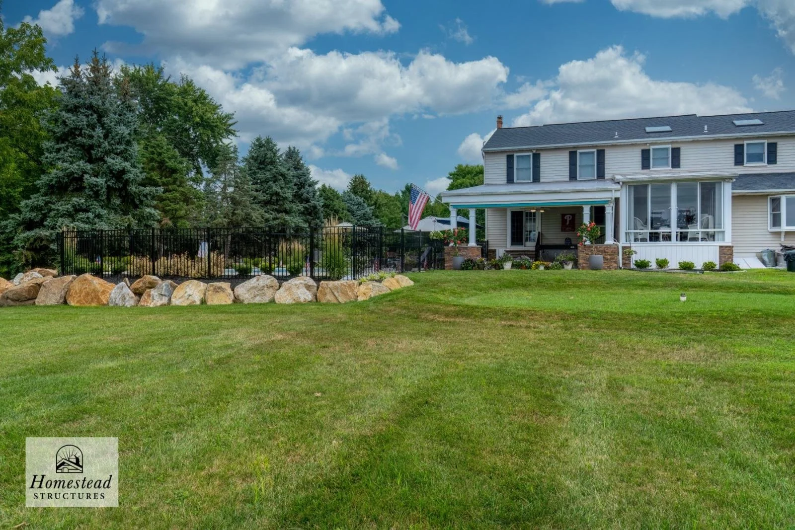 A large two-story house with a front porch decorated with potted plants and a Pennsylvania flag. The house is painted off-white with black shutters and a dark gray roof. There is a well-maintained lawn with a stone border and a wooded backyard with t