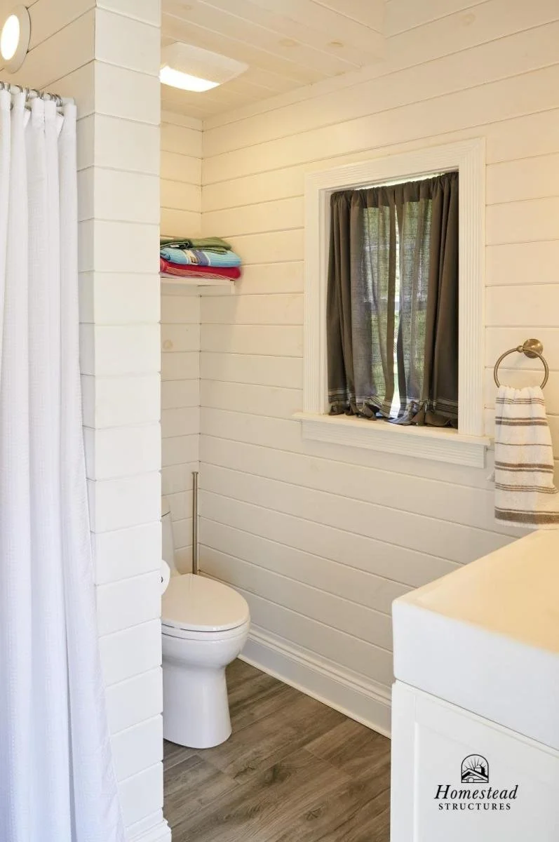 A cozy bathroom featuring white shiplap walls, a toilet beside a window with dark curtains, a shelf with folded towels, a vanity with a towel ring, and a shower with a white curtain.