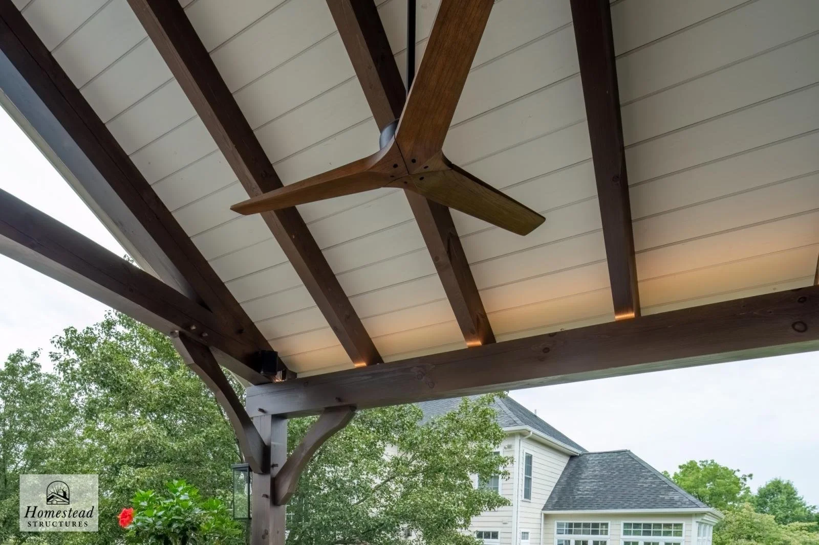 View of a wooden porch ceiling with exposed beams, a ceiling fan, and part of a house with trees in the background.