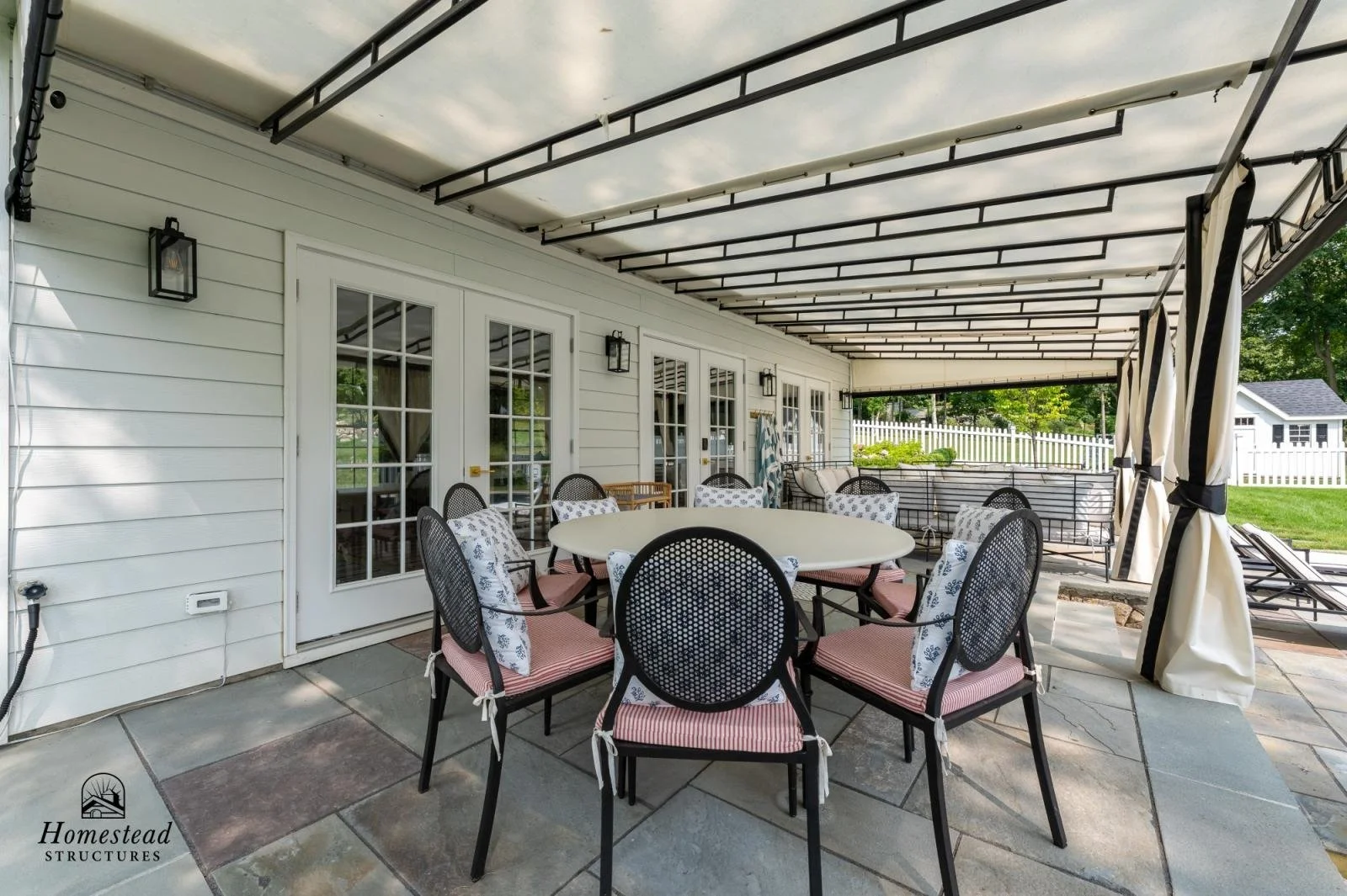 Covered outdoor patio with a round dining table, eight chairs with red and white striped cushions, black metal frames, and floral pillows. Glass-paneled doors on the house wall, lantern-style wall lights, and a white fence and lawn in the background.