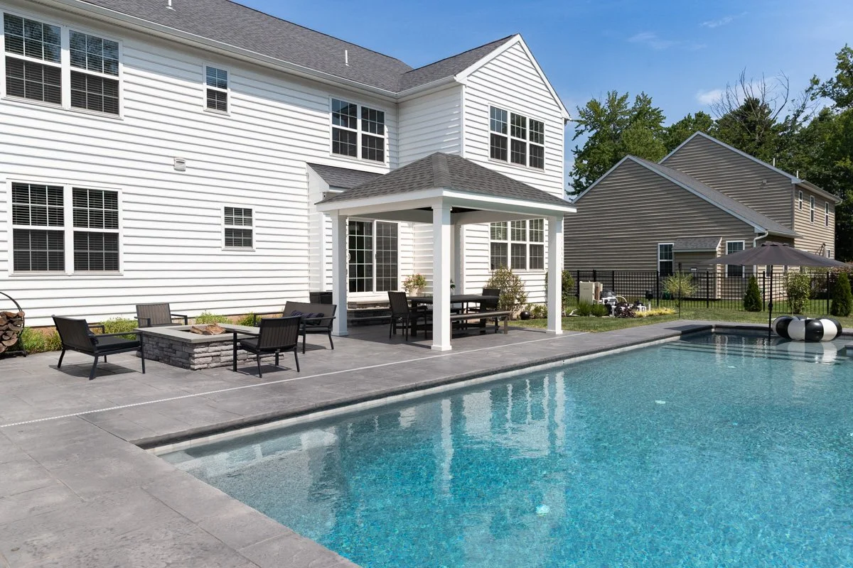Backyard with a swimming pool, patio furniture, and a gazebo next to a white house on a sunny day.