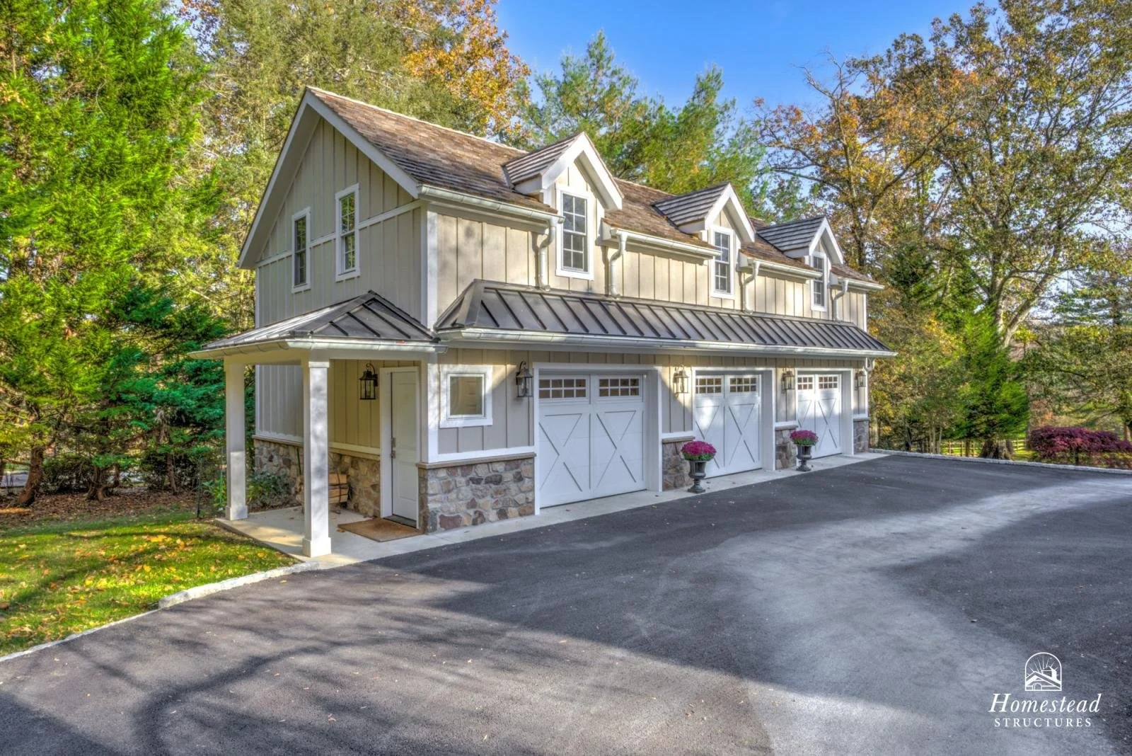 A two-story house with beige siding, stone accents at the base, multiple dormer windows on the roof, and a triple garage door, surrounded by trees with autumn foliage.