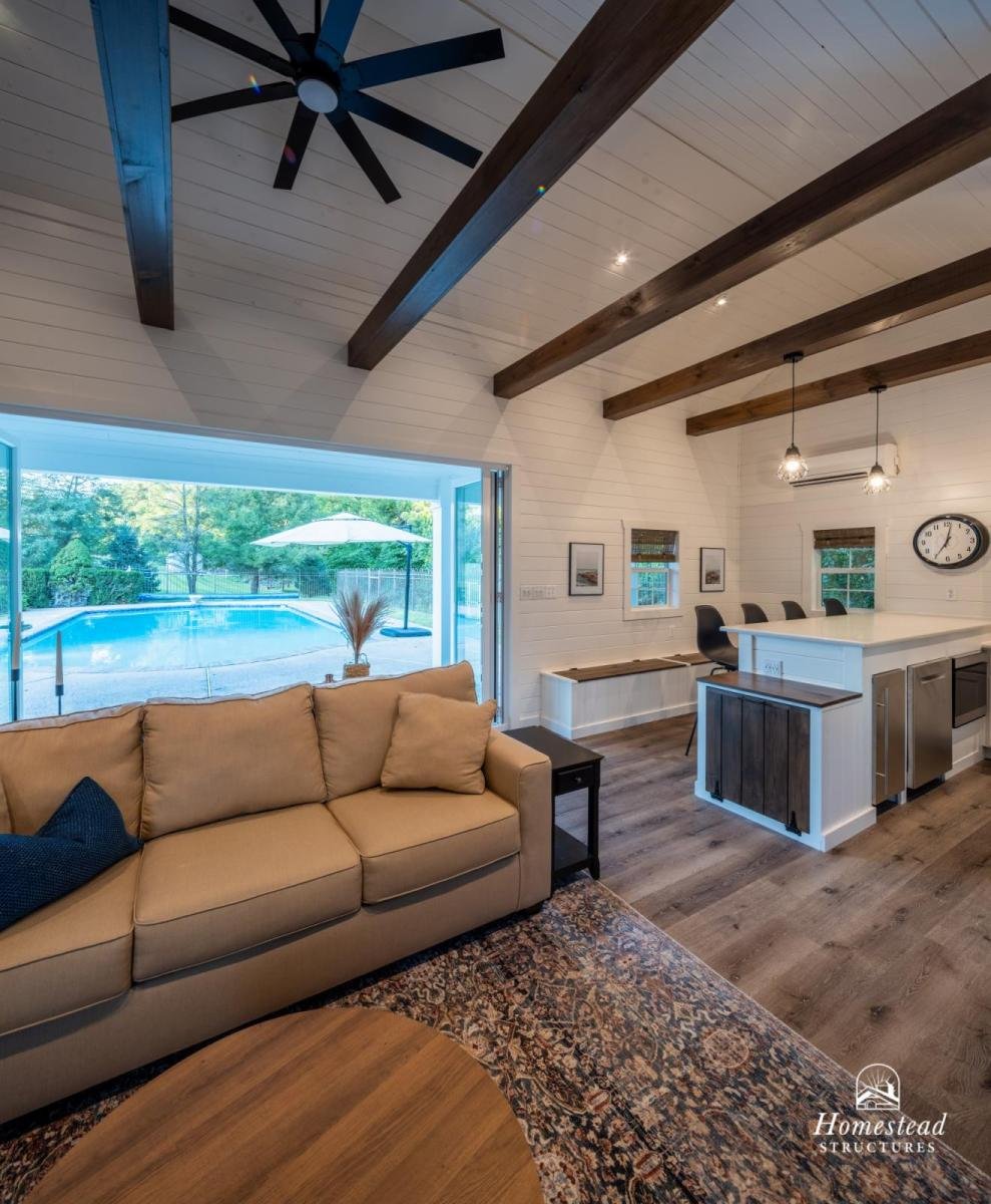 Living room with beige sofa, black side table, wooden coffee table, area rug, and large glass doors opening to a backyard with a pool and umbrella. Modern kitchen with white cabinets, bar stools, black clock, and ceiling fan with wood beams.
