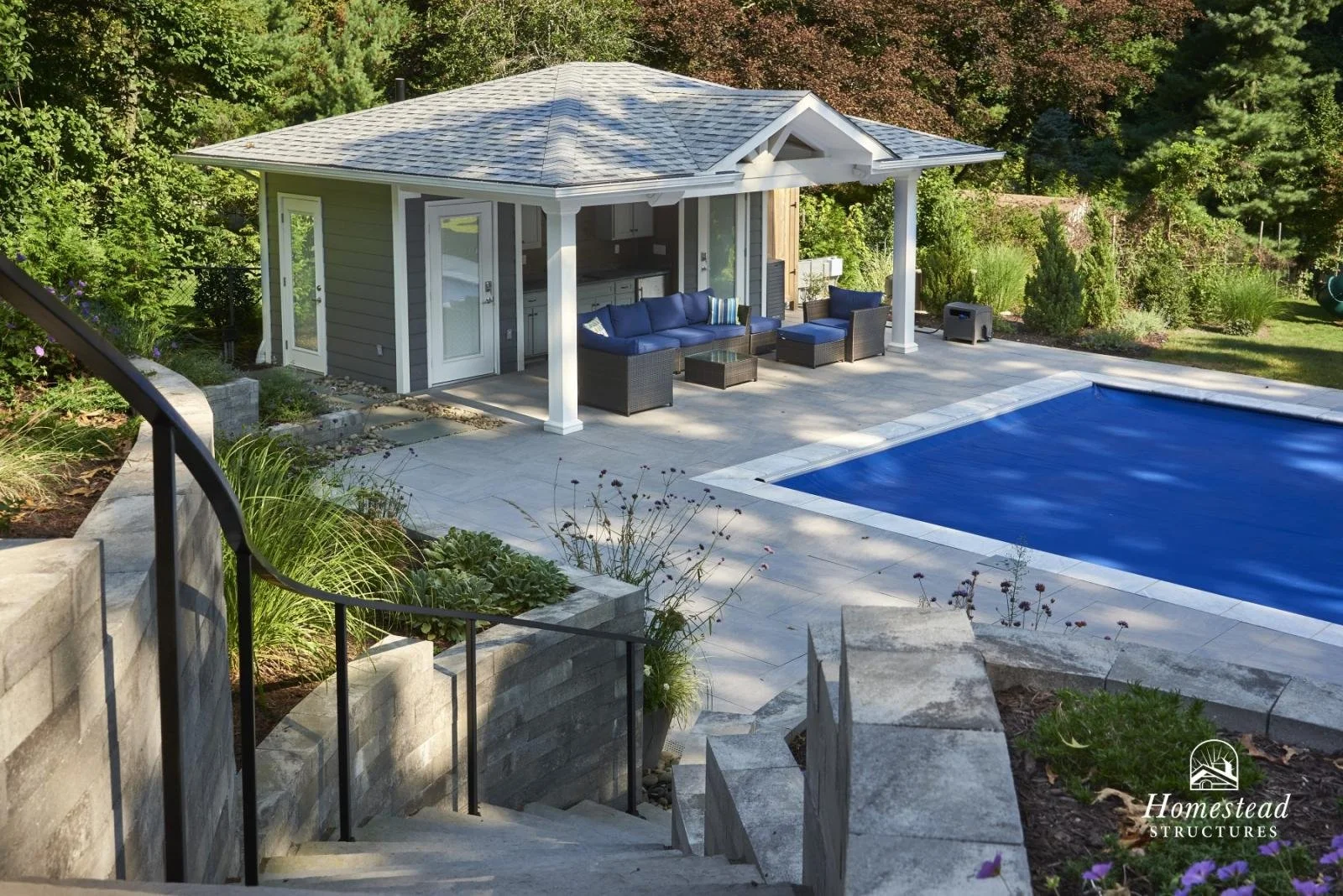 A backyard with a swimming pool, outdoor seating area under a small pavilion, and surrounding greenery.