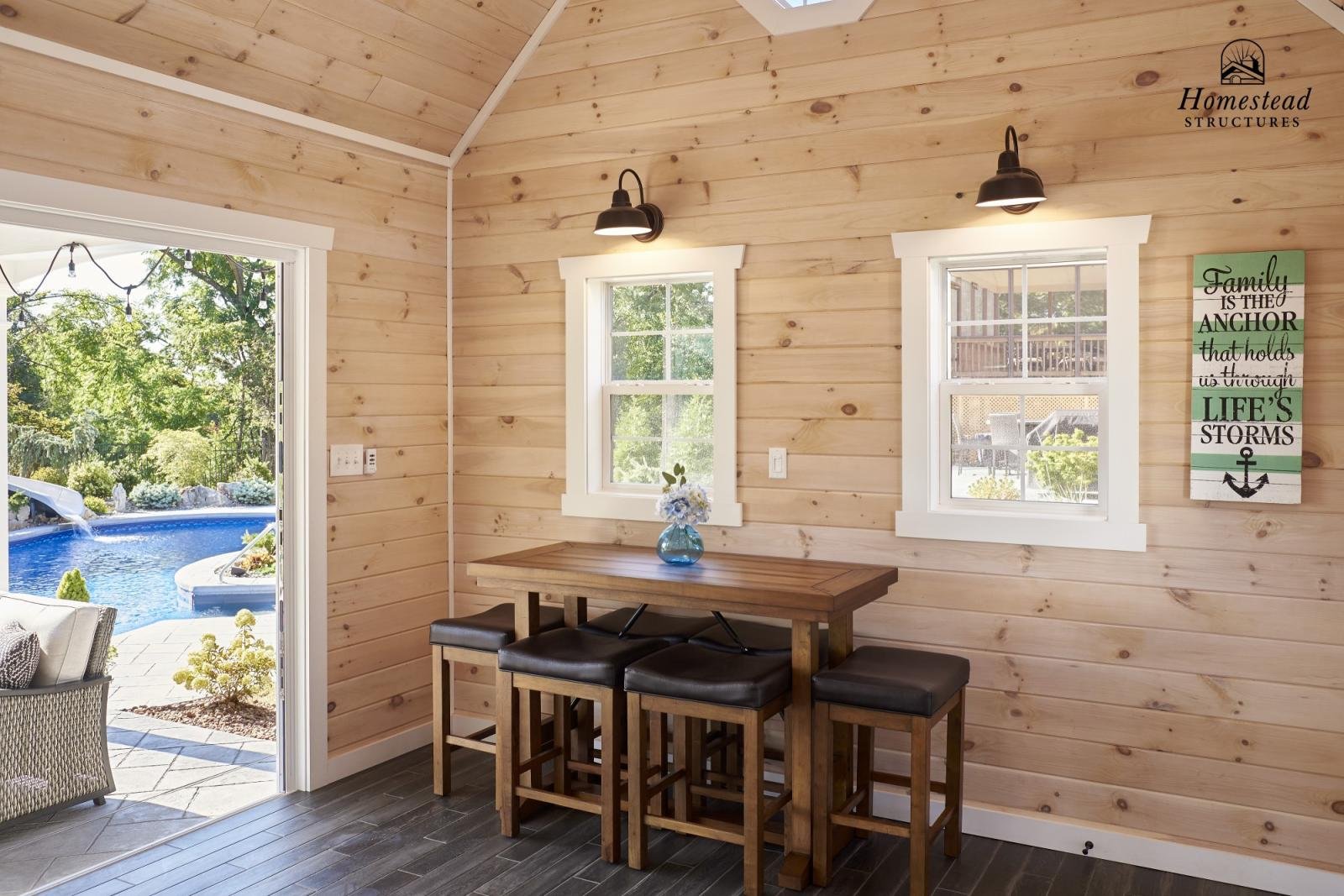 Interior of a small wooden room with two windows, a wooden table with four black-cushioned stools, a vase with flowers, wall-mounted lights, a door leading outside showing a pool, and a decorative sign about family and life's storms.
