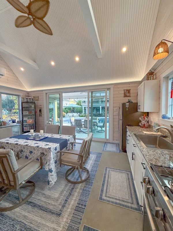 Bright kitchen and dining area with a table covered in a floral tablecloth and four chairs, a sliding glass door leading to an outdoor patio, kitchen countertops with appliances, and a ceiling fan with leaf-shaped blades.