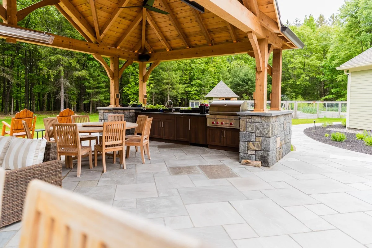 Outdoor patio with wooden pergola, dining table with chairs, outdoor kitchen area with grill, surrounded by lush green trees.