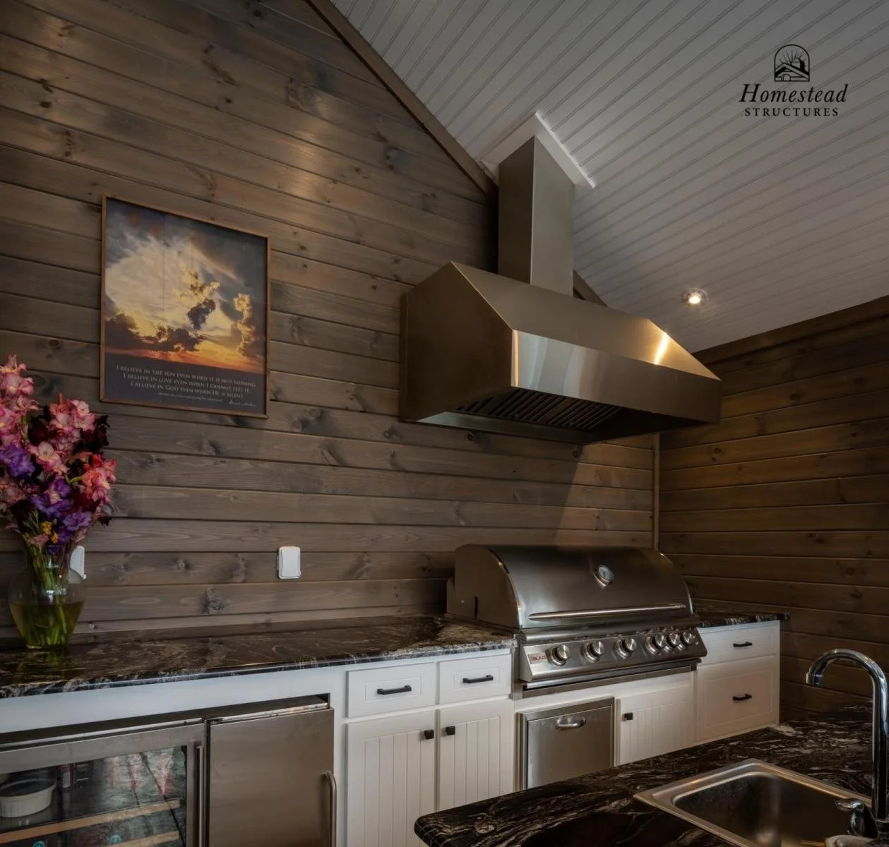 Kitchen with wooden panel walls, stainless steel range hood and grill, white cabinets, black granite countertop, a small wine fridge, a pink and purple flower arrangement on the counter, and framed sunset sky picture on the wall.
