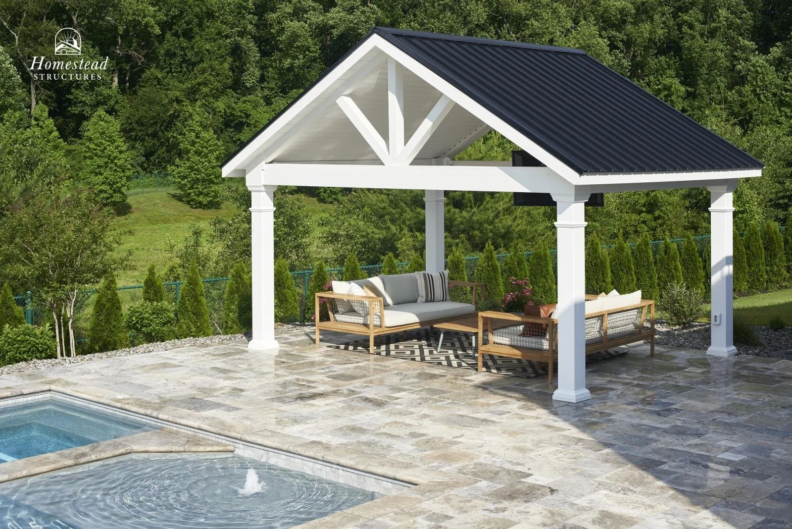 A backyard patio area featuring a white pergola with a black metal roof. Underneath, there are outdoor couches and chairs with cushions, and a patterned rug. Adjacent is a swimming pool with a small fountain, surrounded by a stone deck and green tree