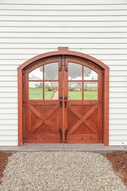 Wooden barn-style double doors with glass windows mounted on white siding, with a gravel pathway in front.