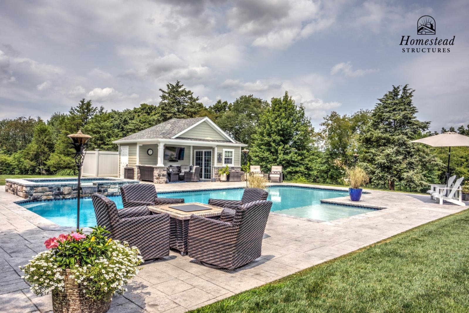 A backyard with a swimming pool and hot tub, outdoor furniture including chairs and an umbrella, and a small building with a TV. Landscaping includes green grass and trees, with a cloudy sky overhead.
