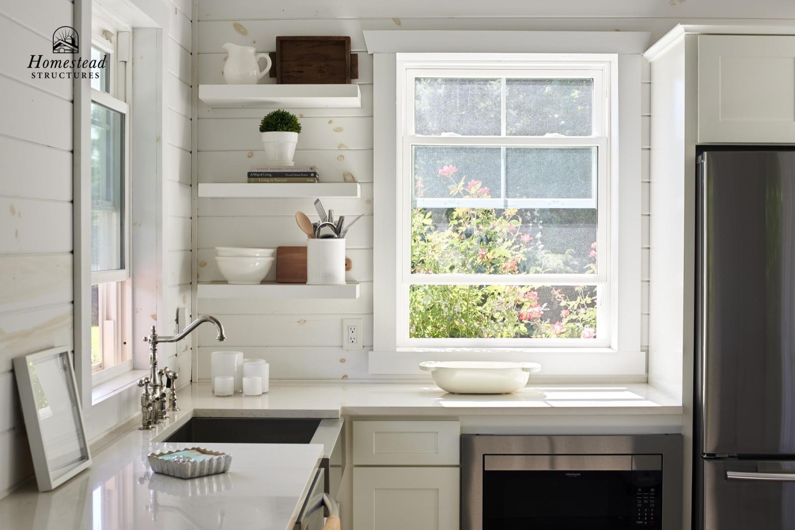 Bright kitchen with white shiplap walls, a window showing green plants outside, white shelves with decorative items, a white countertop with a bowl and candles, a stainless steel refrigerator, and a microwave.