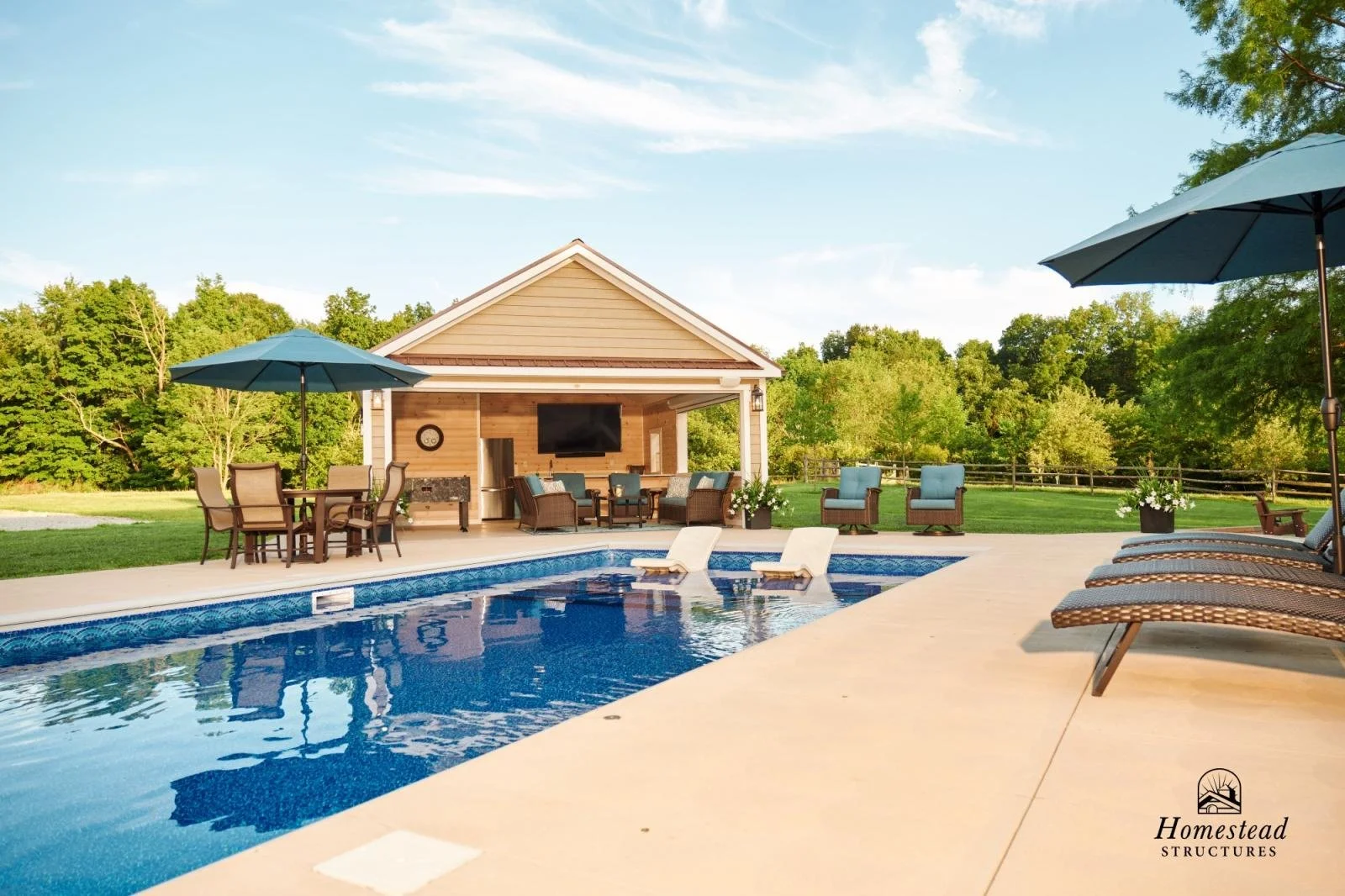 An outdoor backyard with a swimming pool, surrounded by patio chairs and umbrellas, a covered lounge area with outdoor furniture, a flat-screen TV, and lush green trees in the background.