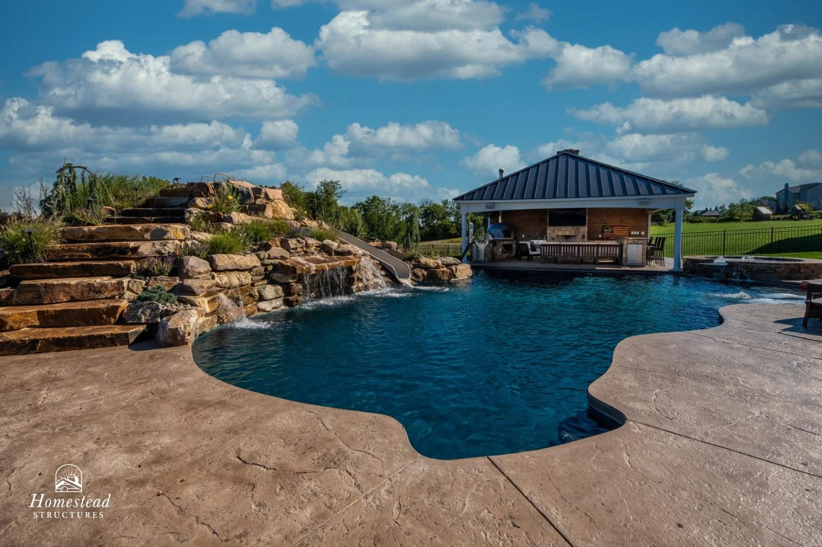 A backyard swimming pool with a rock waterfall feature, surrounded by a textured concrete deck. There is a covered outdoor kitchen and seating area in the background, with a grill and barbecue. The sky is blue with scattered white clouds, and there i