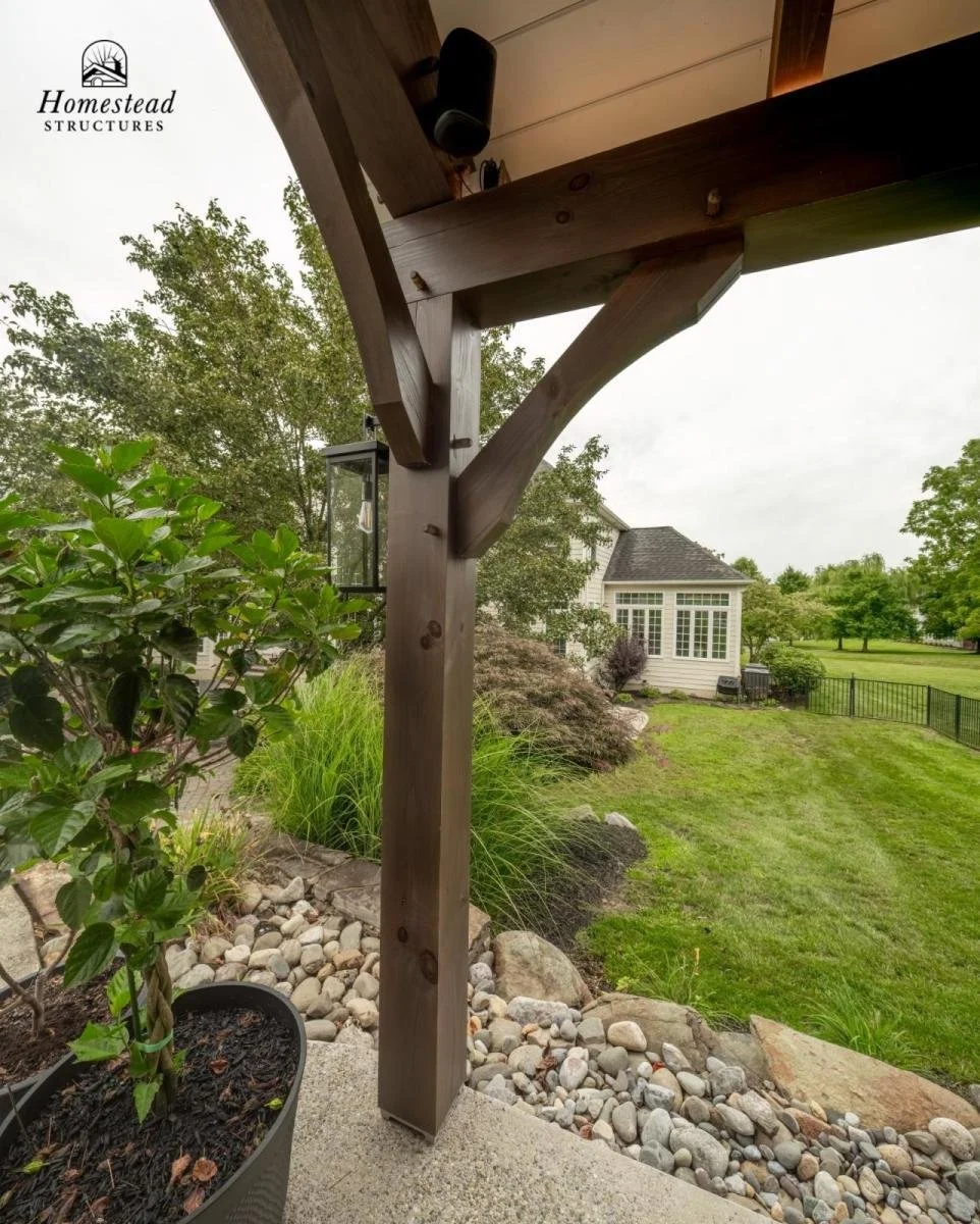 Close-up of a wooden support beam and bracket under an outdoor porch roof, with a landscape of a lawn, shrubs, and a house in the background.