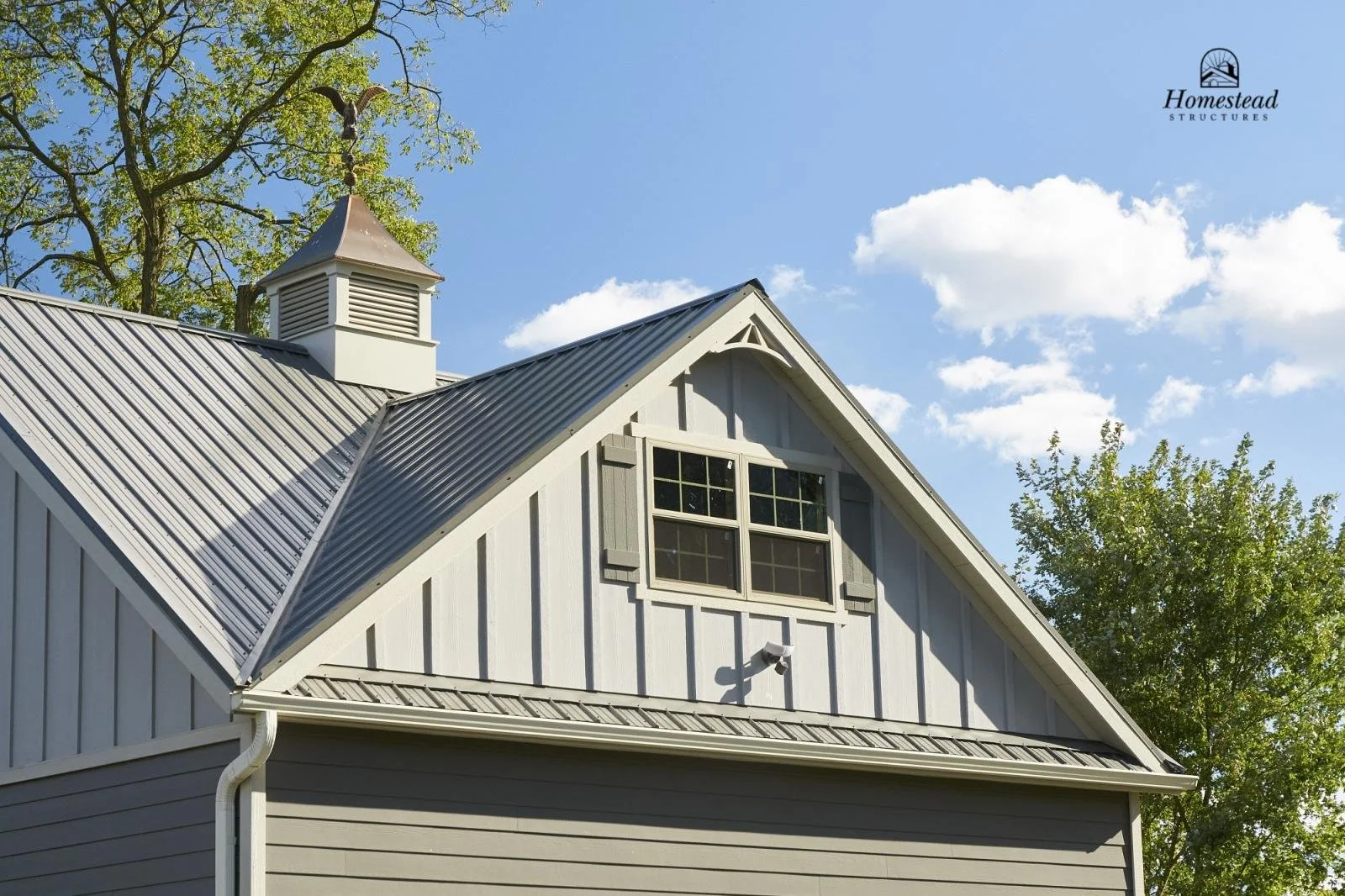 A house with gray siding, a metal roof, and a small dormer window with shutters. There is a cupola at the peak of the roof with a weather vane. Green trees and a blue sky with clouds are in the background.