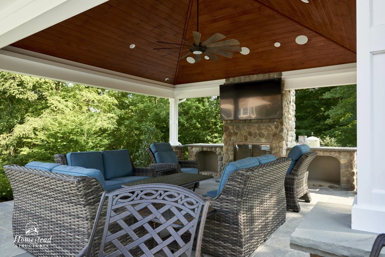 Covered outdoor patio with wicker chairs, blue cushions, a stone fireplace, a mounted flat-screen TV, and a wooden ceiling fan, surrounded by greenery.