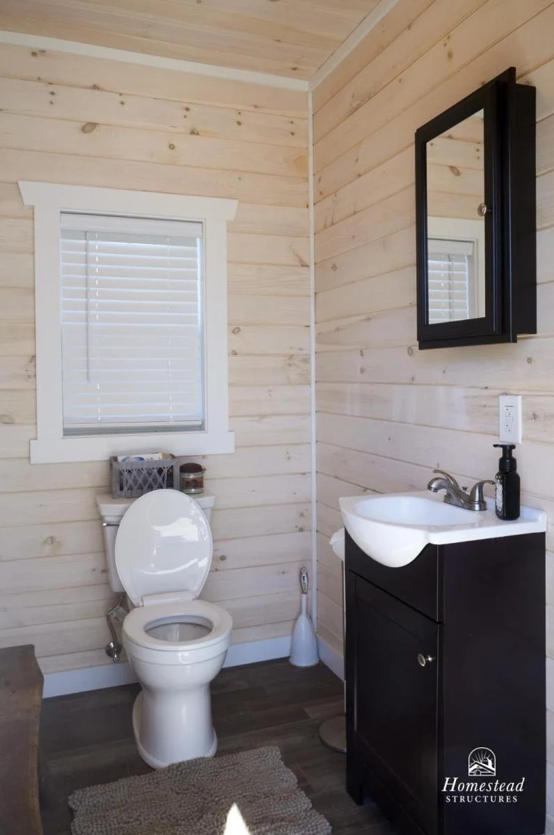 Small bathroom with light-colored wooden walls, a white toilet, a window with blinds, a black cabinet with a white sink, a mirror cabinet, a soap dispenser, a toilet brush, a small rug, and a window.
