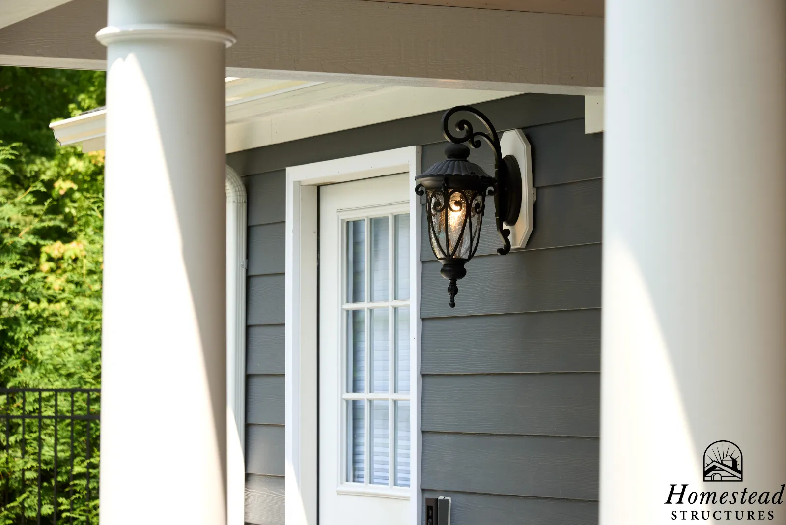 Exterior of a house with gray siding, white trim, a window with white grid, a black outdoor lantern, and a white column partially visible.