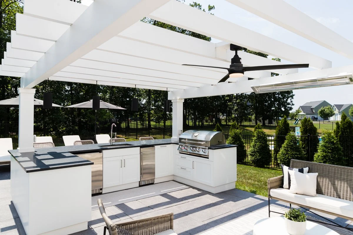 Outdoor kitchen with a white pergola, grill, sink, cabinets, ceiling fan, and seating area with chairs, a sofa, and an outdoor rug in a backyard with trees and houses in the background.